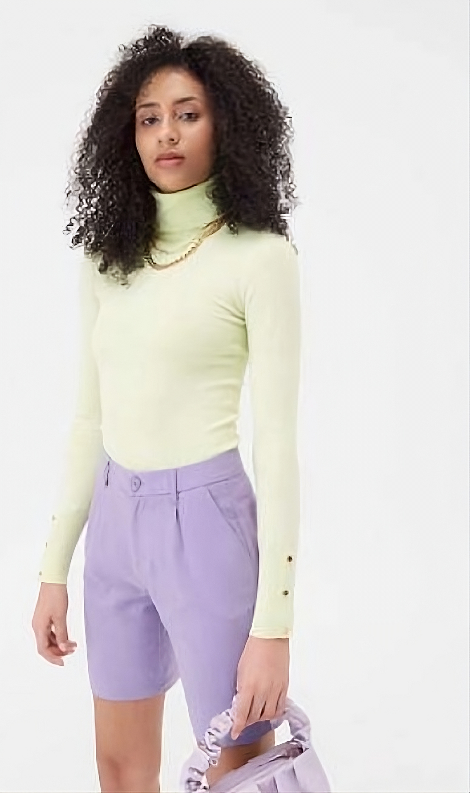 uma moça com uma blusa verde claro de gola alta e manga longa, e um short da cor ciano, e segurando uma bolsa da mesma cor